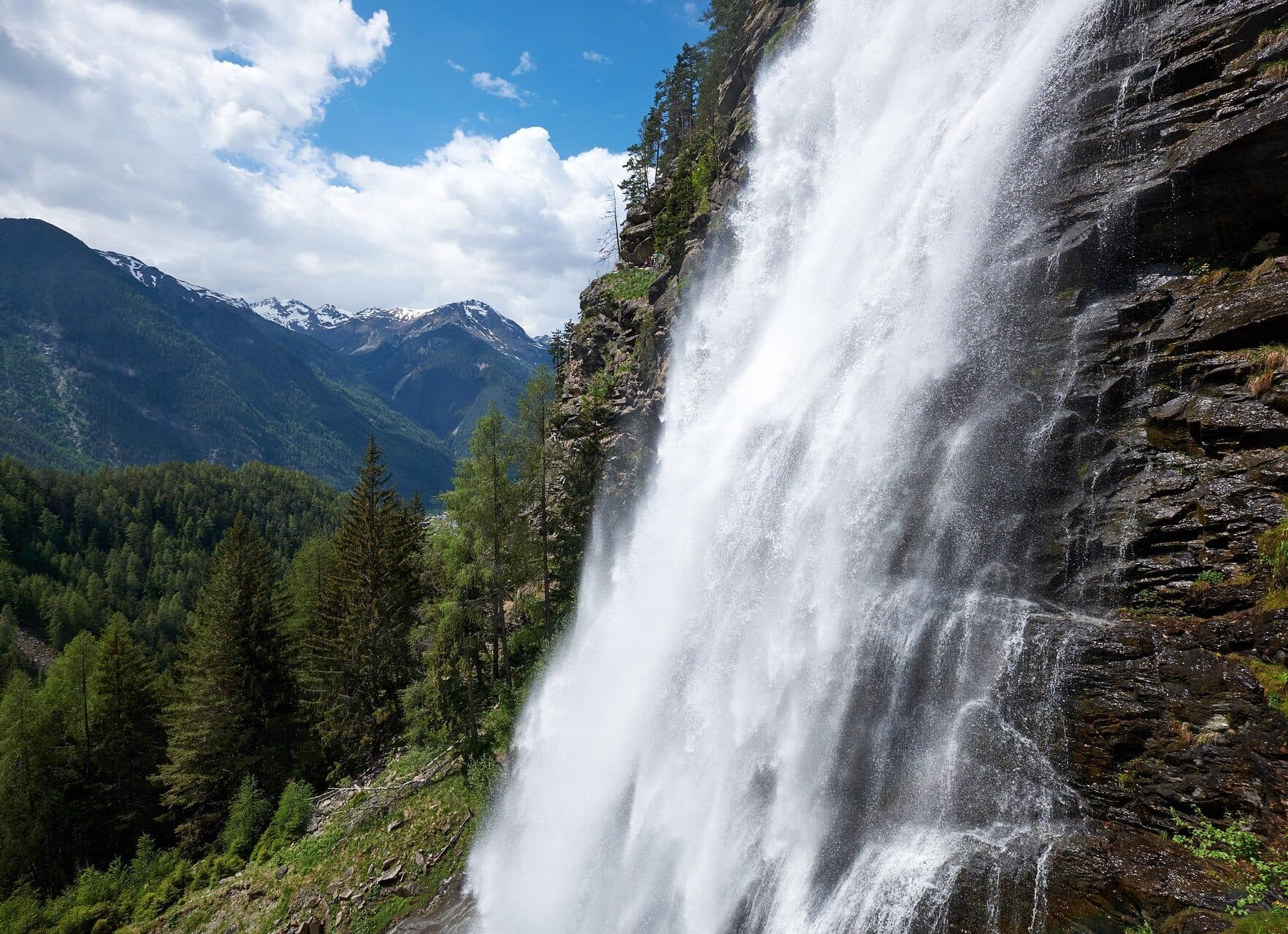 Ein Wasserfall stürzt über eine felsige Klippe mit bewaldeten Bergen und einem teilweise bewölkten Himmel im Hintergrund.