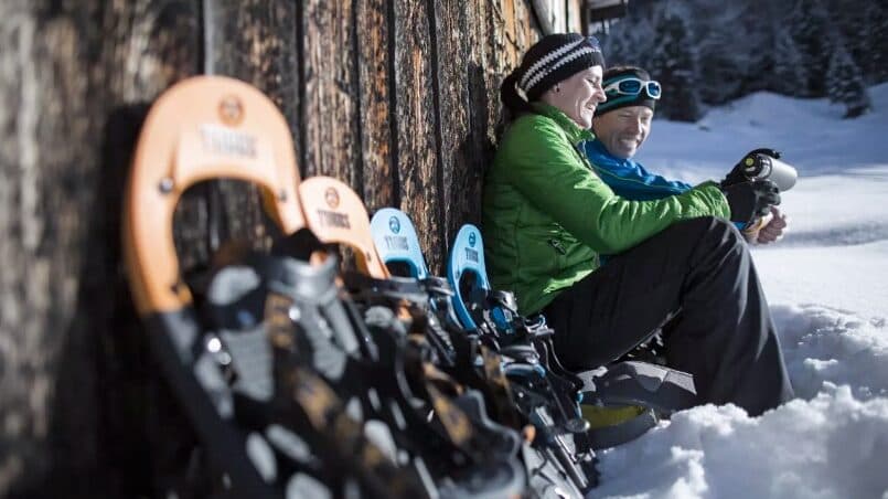 Zwei Personen in Winterkleidung sitzen an einer Holzwand im Schnee und halten Tassen in der Hand. Im Vordergrund sind Schneeschuhe aufgereiht.
