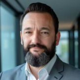 Мужчина с темными волосами и бородой, одетый в серый костюм и белую рубашку, стоит в помещении перед большими окнами.