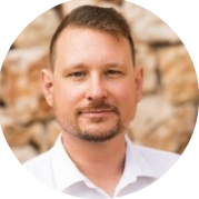 Мужчина с короткими светло-каштановыми волосами, подстриженной бородой и усами, в белой клетчатой рубашке стоит на фоне каменной стены.