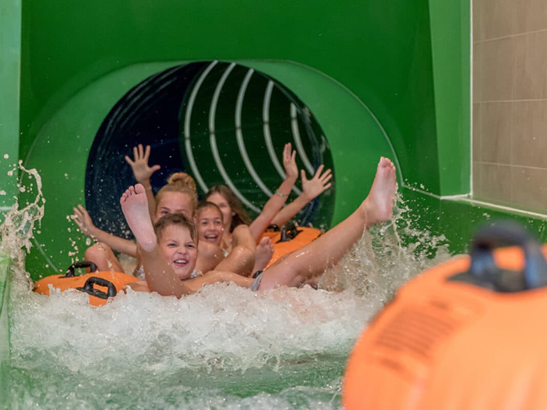 Vier Kinder fahren gemeinsam auf einem orangefarbenen Floß und spritzen mit erhobenen Händen und Beinen aus einer grünen Indoor-Wasserrutsche.