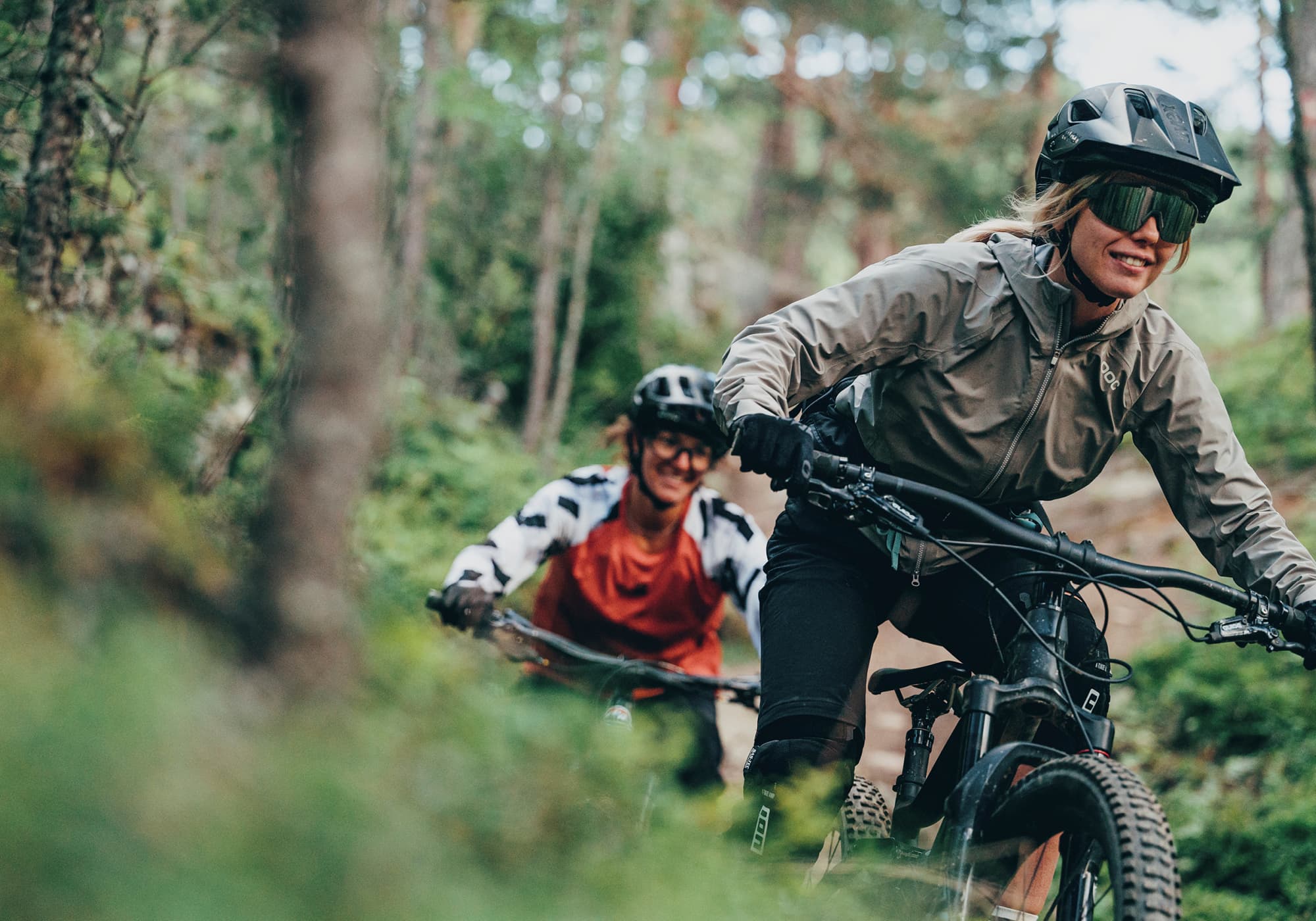 Due persone in mountain bike su un sentiero forestale, entrambe con casco e occhiali da sole. La persona davanti sorride, seguita da vicino da un altro ciclista.