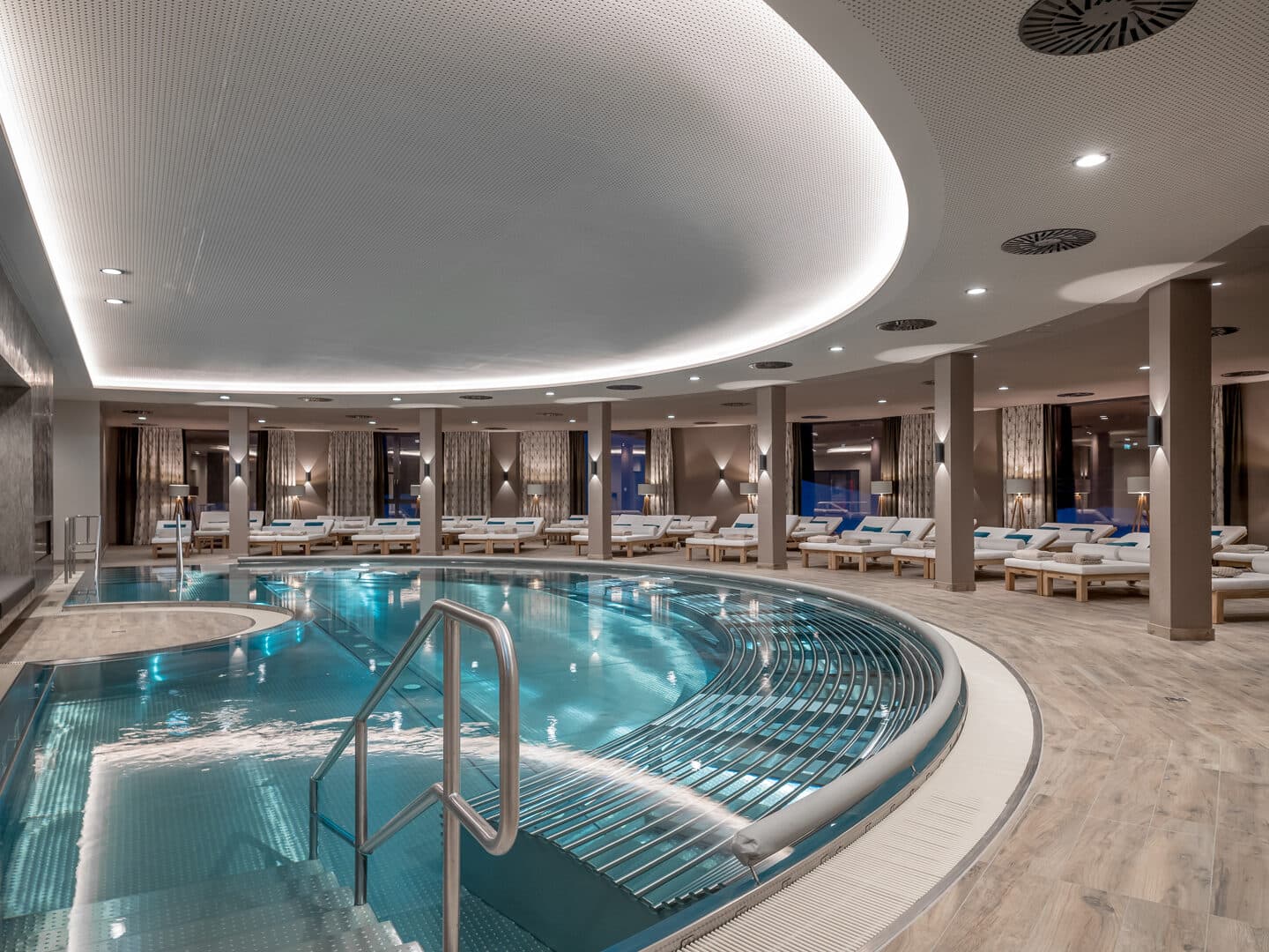 Wellnessbereich im Innenbereich mit einem großen runden Pool, Stufen, die ins Wasser führen, Liegestühlen am Rand und sanfter Beleuchtung über dem Kopf.