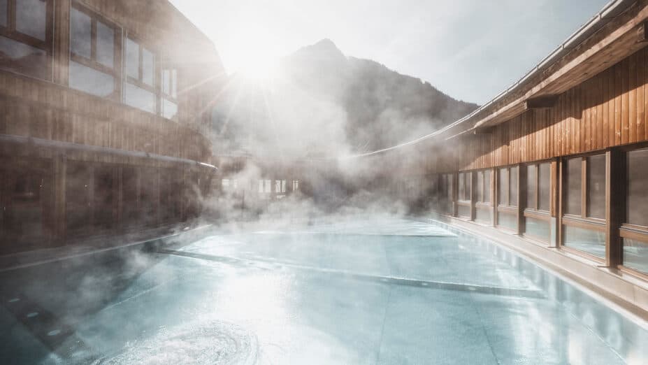Ein dampfendes Außenbecken, umgeben von Holzgebäuden, mit Sonnenlicht, das auf einen Berg im Hintergrund scheint.