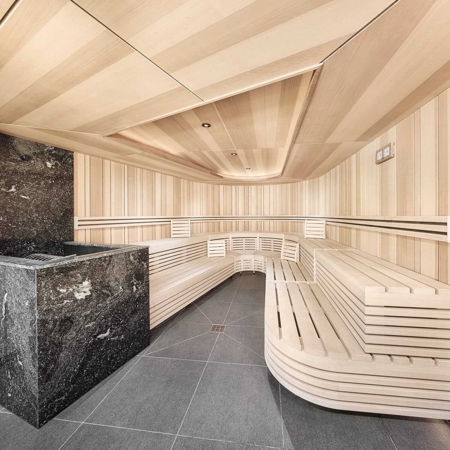 Moderne Holzsauna mit gestuften Bänken, heller Holzverkleidung und einem Saunaofen aus schwarzem Stein auf einem gefliesten Boden.