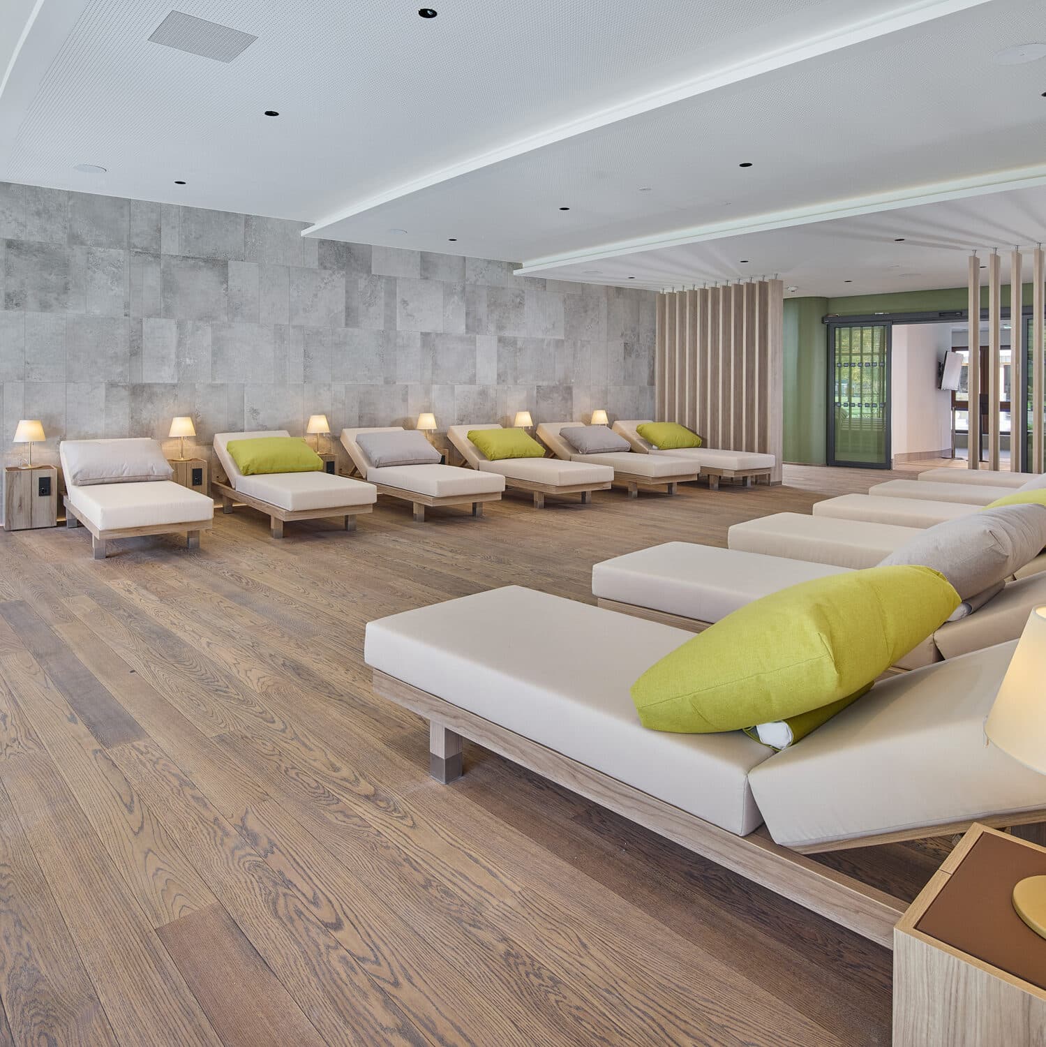 Eine moderne Entspannungslounge mit mehreren beigen Chaiselounges, grünen Akzentkissen, kleinen Beistelltischen und sanfter Beleuchtung auf einem Holzboden.