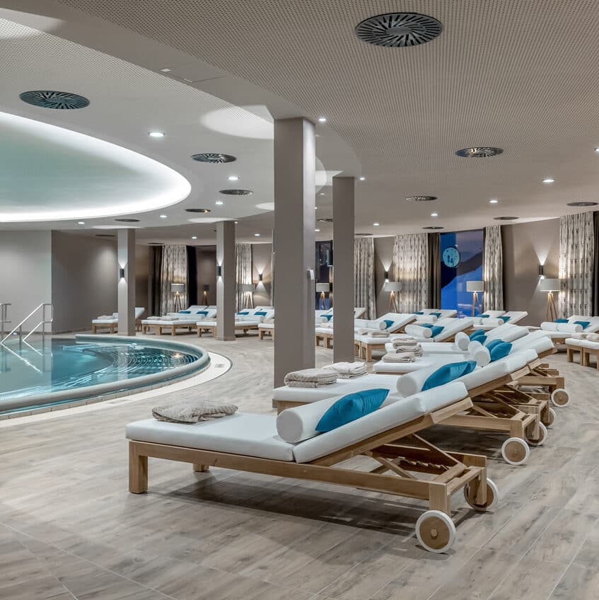 Wellnessbereich im Innenbereich mit mehreren hölzernen Liegestühlen mit weißen und blauen Kissen, die neben einem runden Pool unter sanfter Deckenbeleuchtung angeordnet sind.