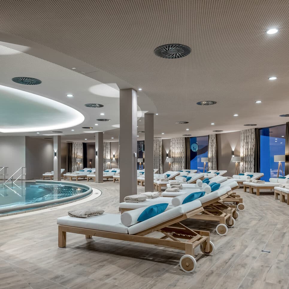 Un'ampia area benessere interna con sedie a sdraio in legno e cuscini blu disposti intorno a una piscina circolare, caratterizzata da un'illuminazione moderna e da finestre a tutta altezza.