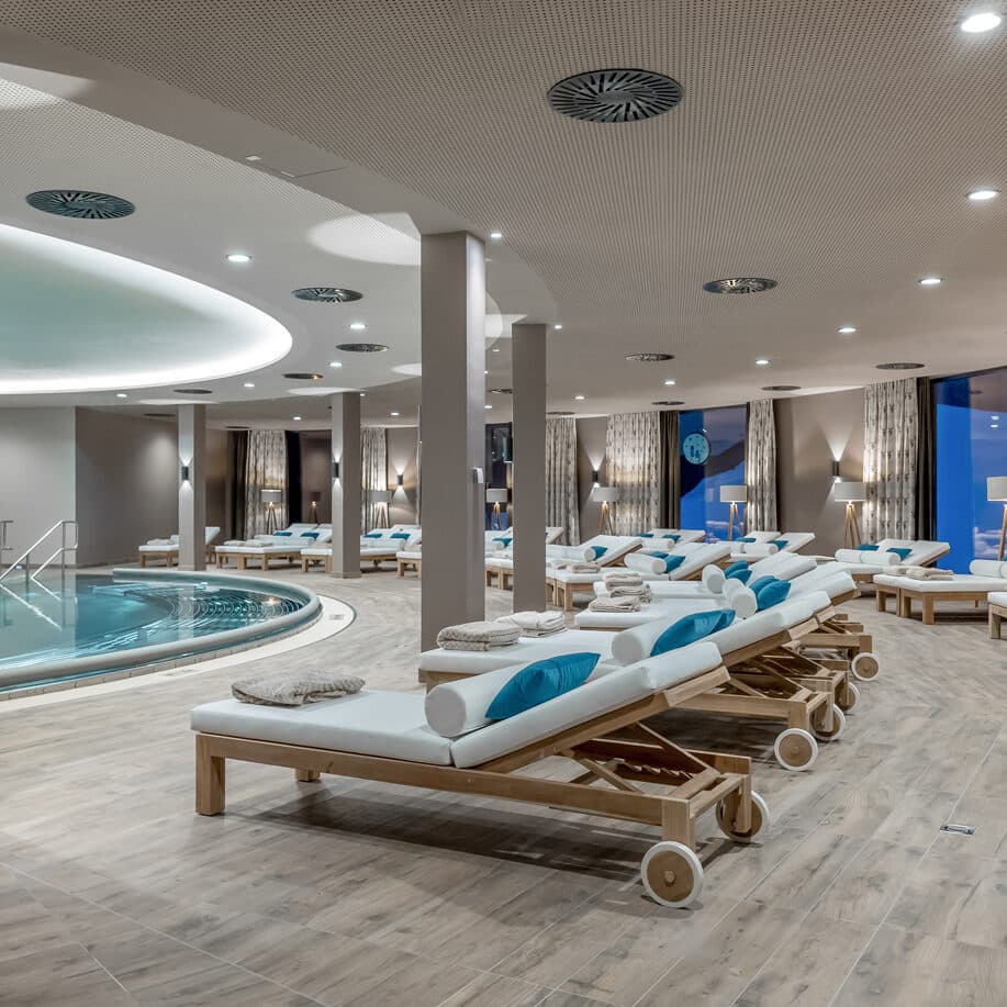Indoor spa area with rows of cushioned lounge chairs, blue pillows, and towels facing a round swimming pool on a wooden floor.