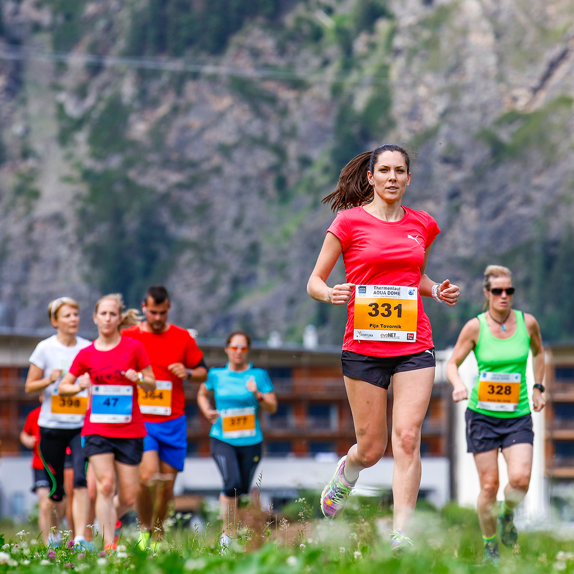 Un gruppo di corridori, ognuno con un pettorale numerato, partecipa a una gara all'aperto con montagne ed edifici sullo sfondo.