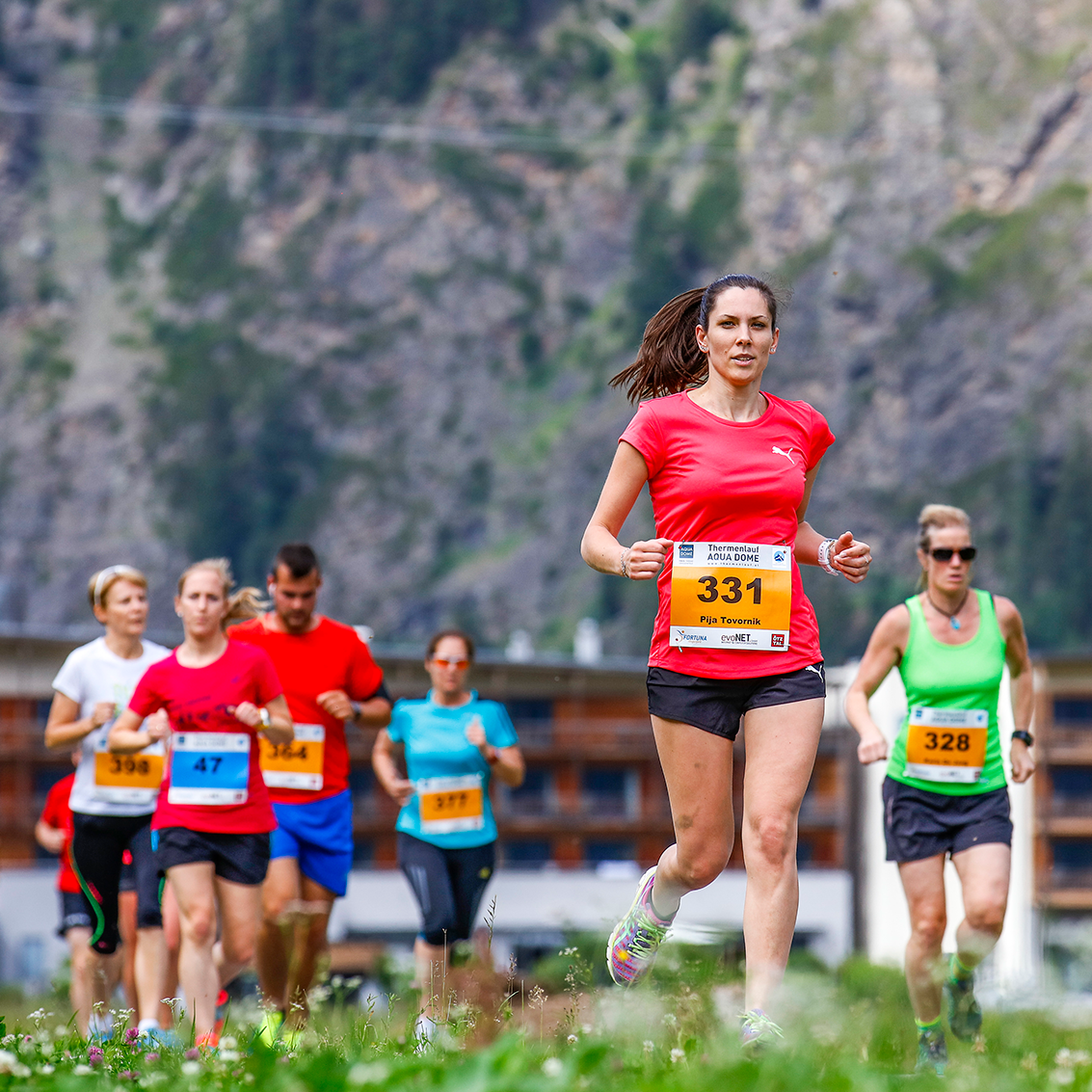 A group of runners participating in a race, with a woman in a red shirt and race bib 331 leading the group outdoors in a mountainous area.