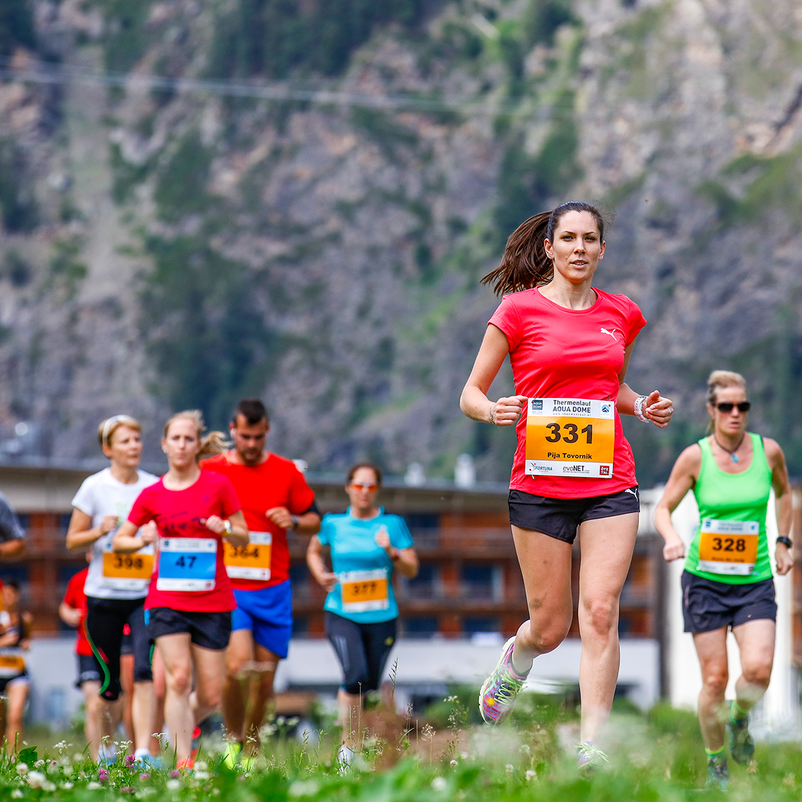 Eine Gruppe von Läufern, die alle Startnummern tragen, nimmt an einem Marathonlauf im Freien teil, mit Bergen und Gebäuden im Hintergrund.