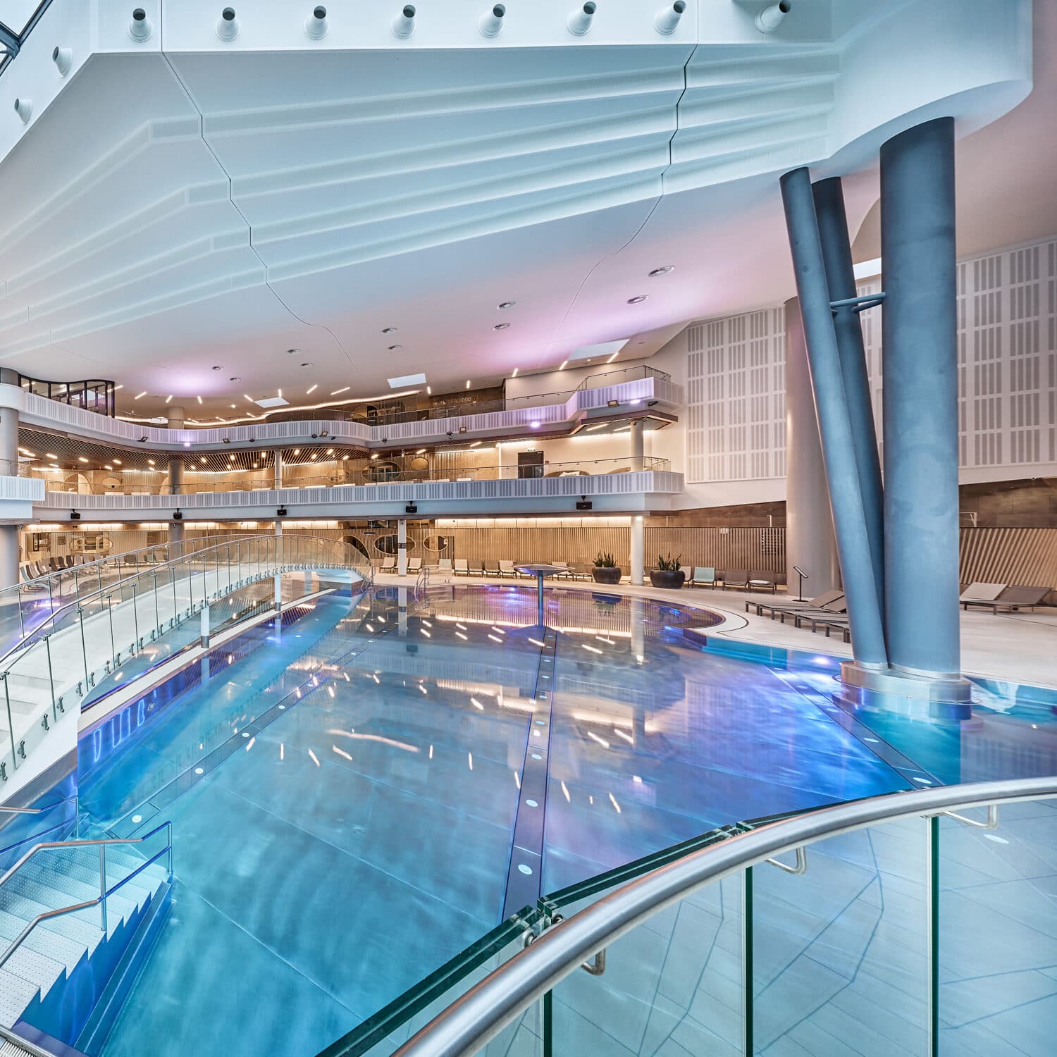 Moderner Hallenbadkomplex mit klarem, blauem Wasser, mehreren Balkonen, Glasgeländern, Liegestühlen und großen Struktursäulen, beleuchtet von Decken- und Akzentlichtern.