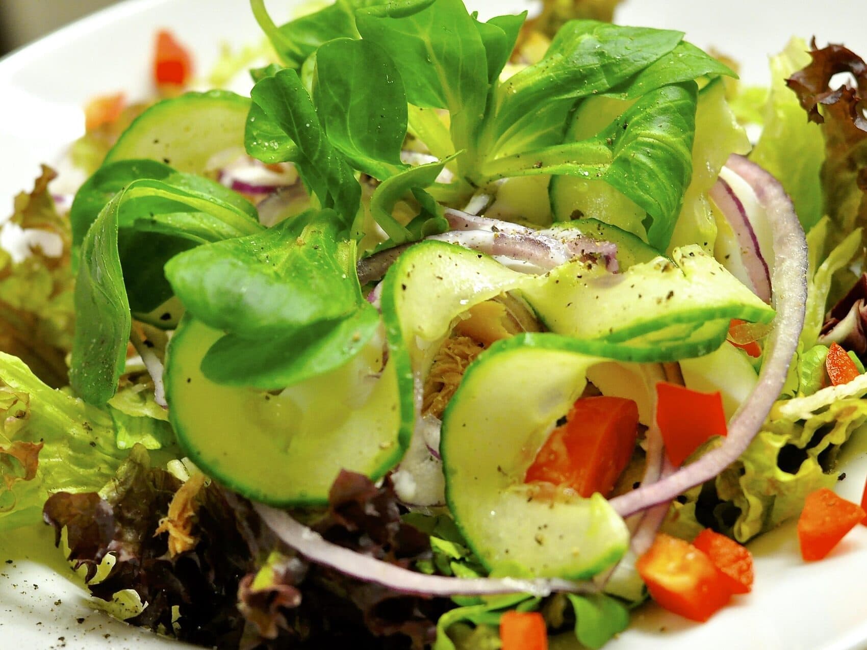 A fresh salad with sliced cucumber, red onion, red bell pepper, leafy greens, and black pepper on a white plate.