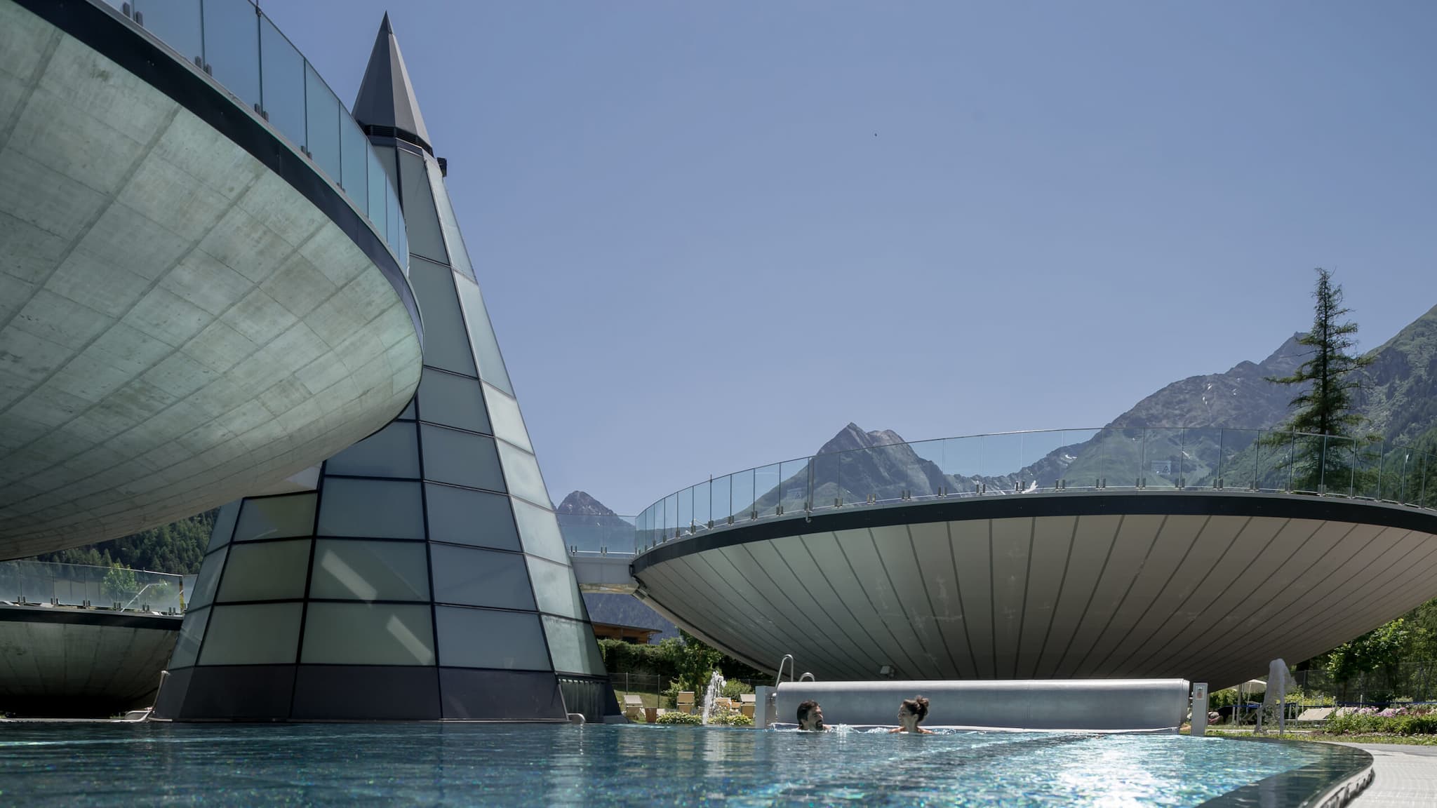 Strutture architettoniche moderne con elementi in vetro e cemento circondano una piscina all'aperto, con le montagne che si stagliano sullo sfondo sotto un cielo limpido.
