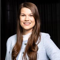 Eine Frau mit langen braunen Haaren trägt einen hellblauen Blazer und ein weißes Oberteil und lächelt in die Kamera vor einem dunklen Hintergrund.