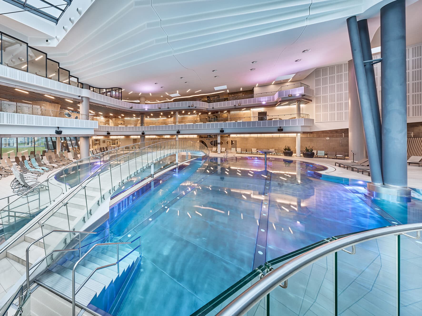 Hallenbad mit klarem Wasser, Glasgeländern, Liegestühlen und mehreren Ebenen. Modernes Design mit weißer Decke und stimmungsvoller Beleuchtung. Keine Personen zu sehen.