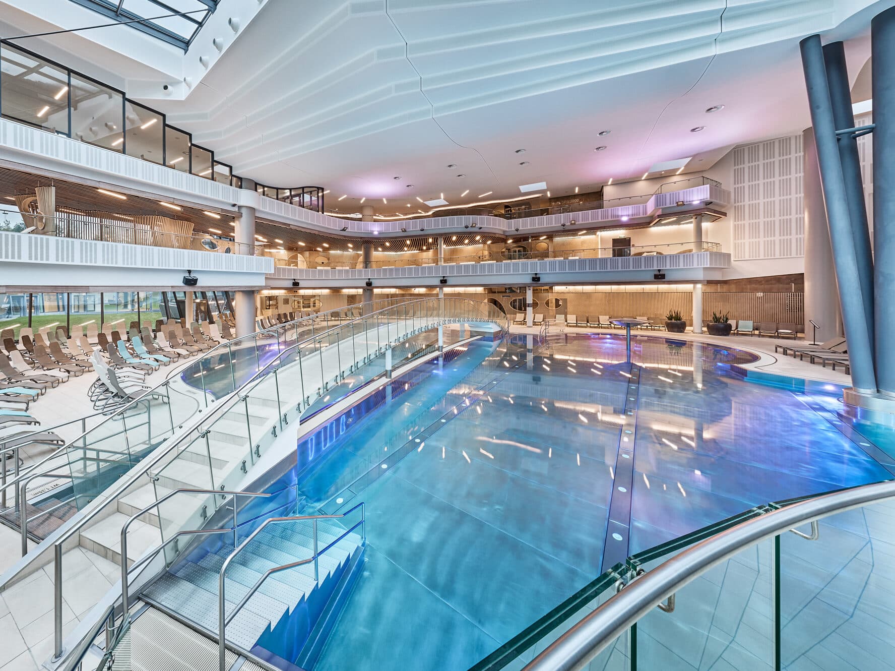 Piscina coperta con acqua cristallina, circondata da sedie a sdraio, ringhiere in vetro e diversi livelli all’interno di una moderna struttura ricreativa.