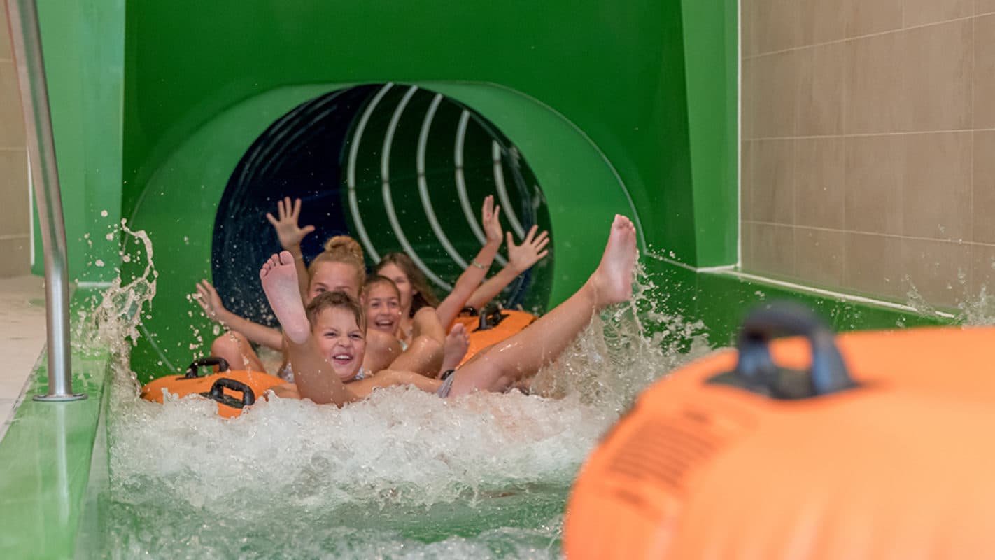 Vier Kinder sitzen gemeinsam auf einem orangefarbenen Schlauchboot, tauchen aus dem grünen Tunnel einer Wasserrutsche auf und springen mit erhobenen Armen ins Becken.