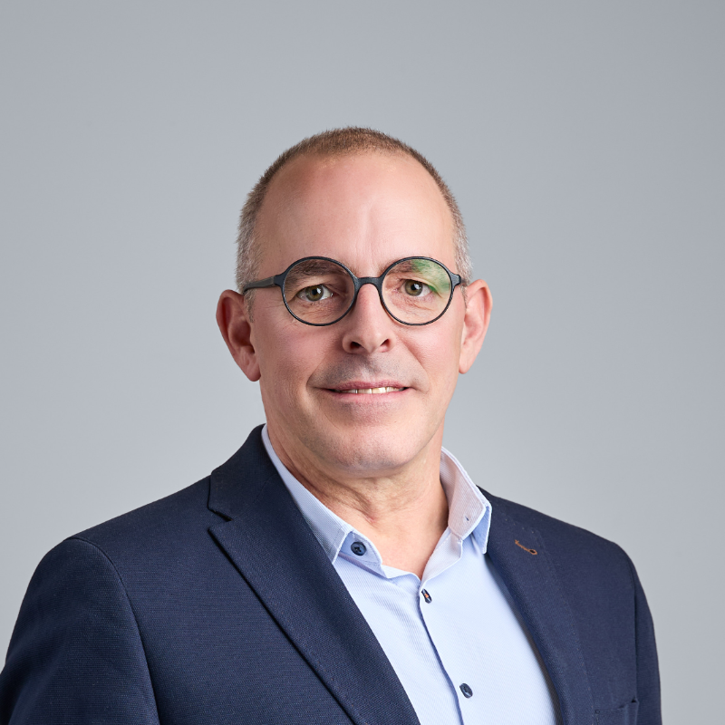 Ein Mann mittleren Alters mit kurzen Haaren und Brille, der einen blauen Blazer und ein hellblaues Hemd mit Knöpfen trägt.