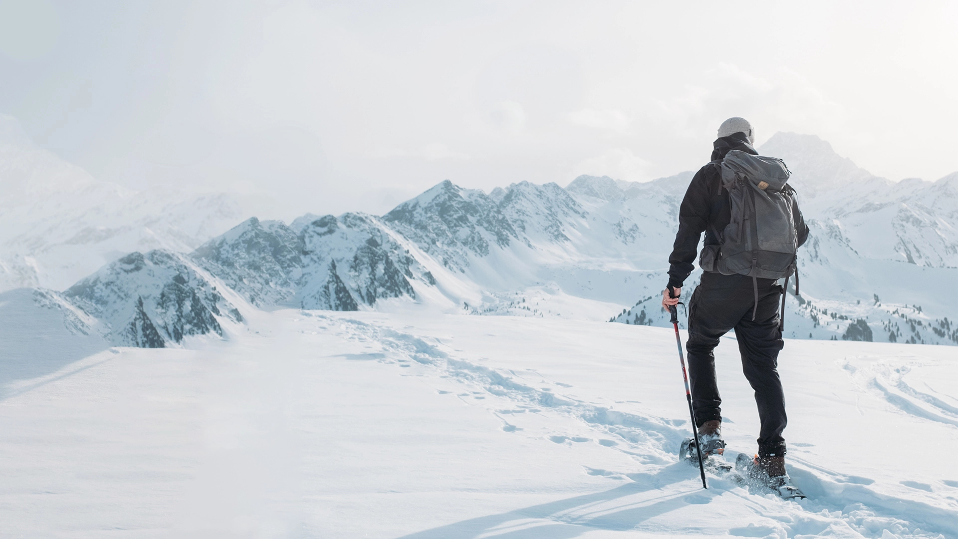 Eine Person in Winterkleidung wandert mit Stöcken durch eine verschneite Landschaft in Richtung der fernen Berge.