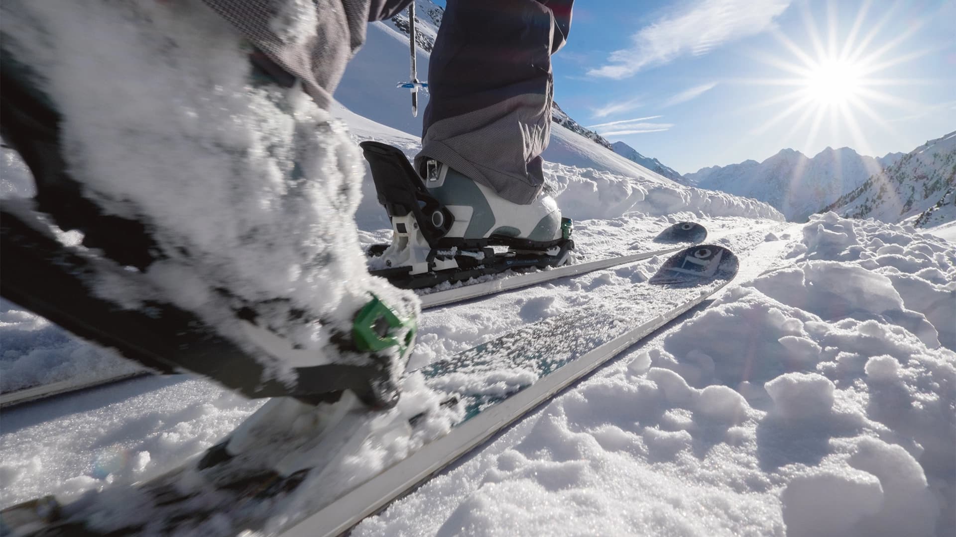Nahaufnahme der Schuhe und Skier eines Skifahrers auf einem schneebedeckten Hang, mit strahlender Sonne und Berggipfeln im Hintergrund.