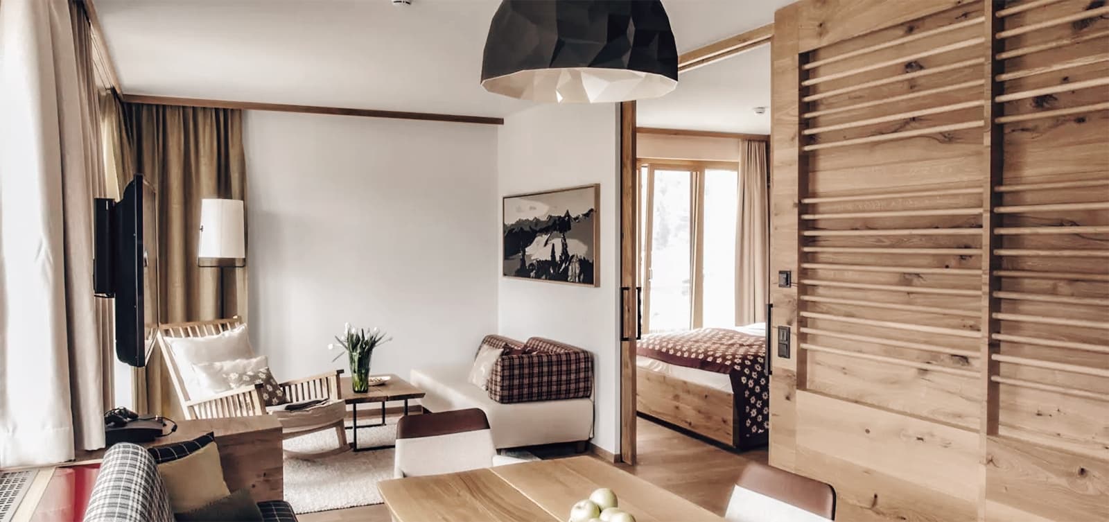 Ein moderner Wohnbereich mit Holzakzenten, einem weißen Sessel, einem kleinen Couchtisch und einem an der Wand montierten Fernseher. Eine teilweise geöffnete Schiebetür gibt den Blick auf ein Schlafzimmer mit großem Fenster frei.