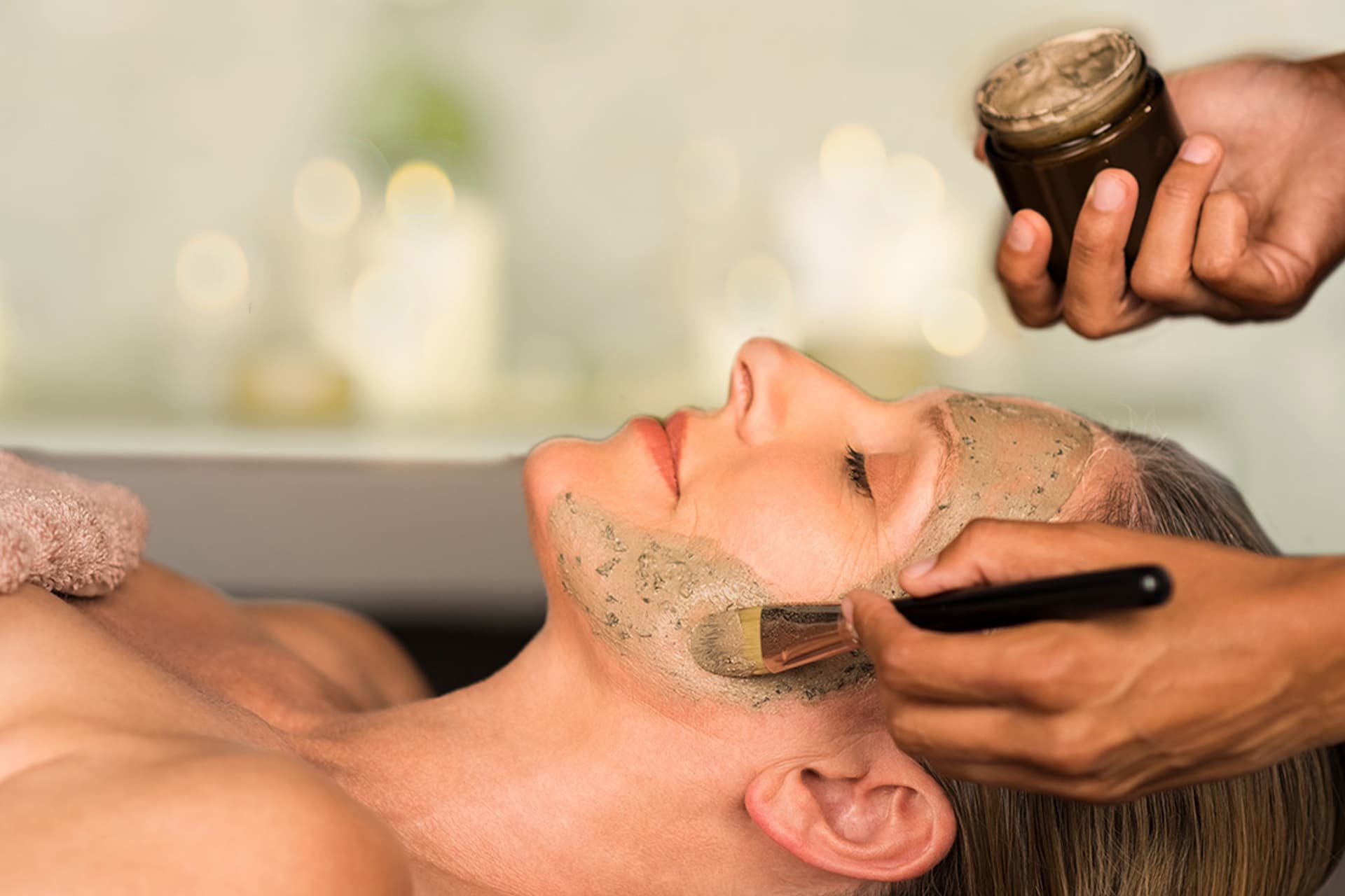 Una persona sdraiata riceve un trattamento viso con un pennello che applica una maschera verde. Un'altra mano tiene un piccolo barattolo di maschera.