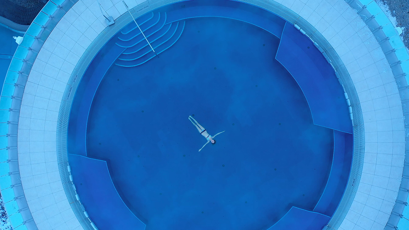 Persona sdraiata sulla schiena in una piscina circolare vuota, vista dall'alto. La piscina ha gradini su un lato ed è circondata da un bordo pavimentato bianco.
