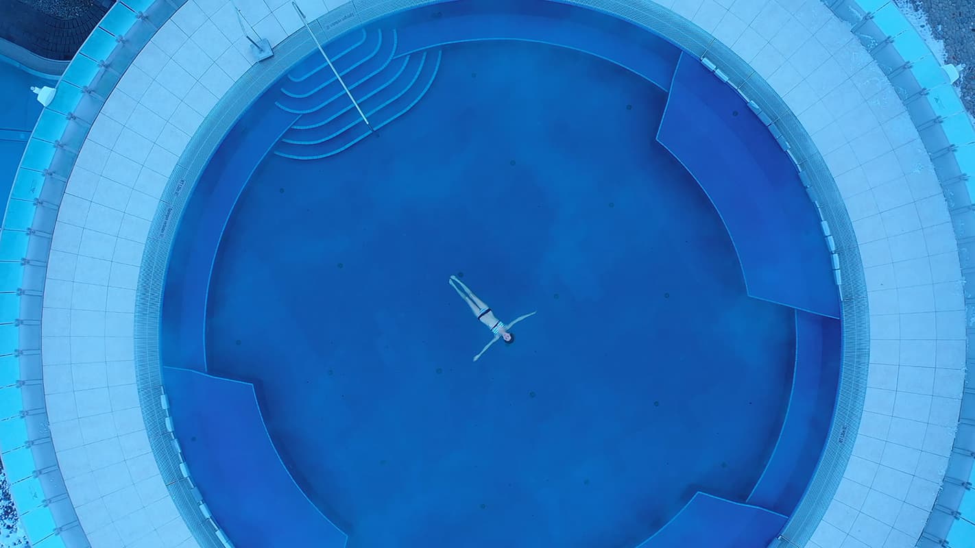 Person floating on their back in an empty, circular blue pool, viewed from above.