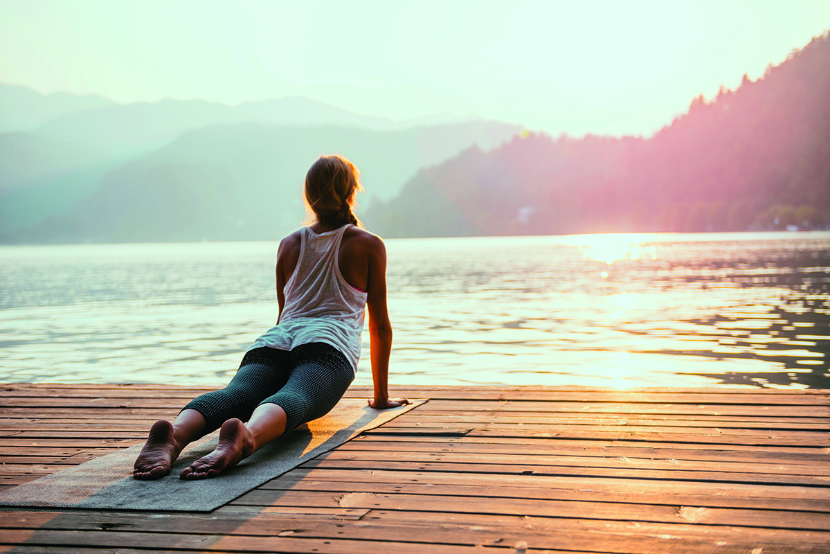 Eine Person praktiziert Yoga bei Sonnenuntergang auf einem Holzsteg an einem See, umgeben von Bergen.