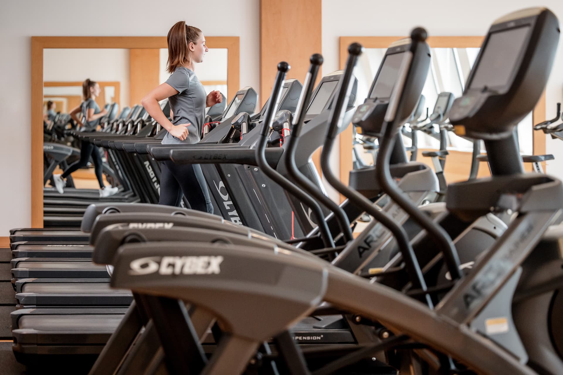 Eine Frau läuft auf einem Laufband in einem Fitnessstudio, umgeben von mehreren anderen Laufbändern. An den Wänden sind große Spiegel zu sehen.
