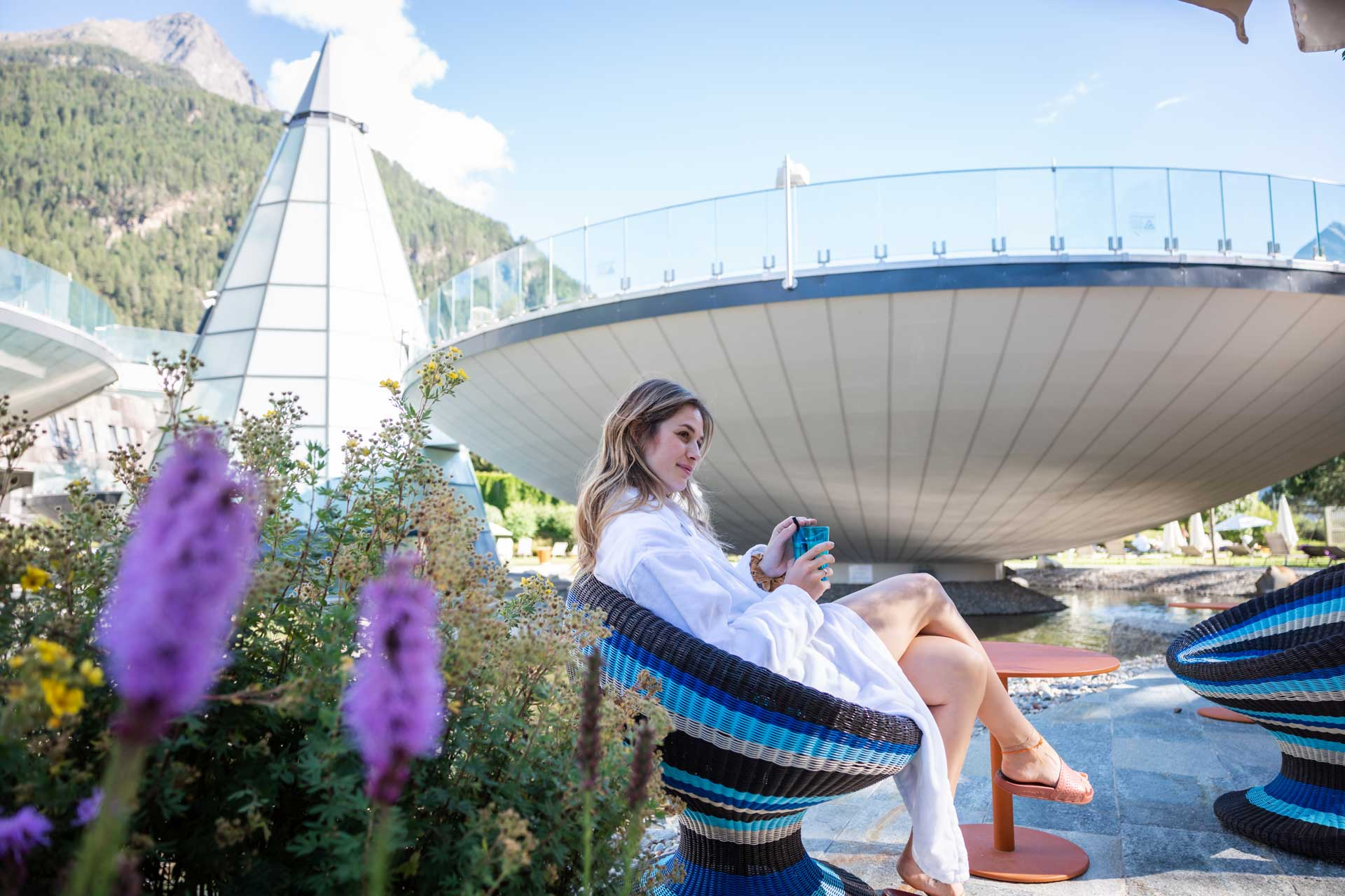 Eine Frau in einem weißen Gewand sitzt auf einem blauen Stuhl und hält eine Tasse in der Hand. Im Freien ist eine moderne Architektur mit Bergen im Hintergrund zu sehen. Im Vordergrund sind Blumen zu sehen.