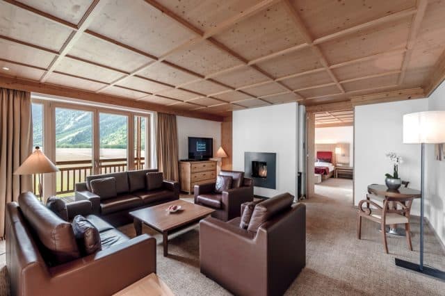 Уютная гостиная с коричневыми кожаными диванами, настенным телевизором и камином. Большие окна открывают живописный вид на горы. Открытый дверной проем ведет в спальню с красным покрывалом.
