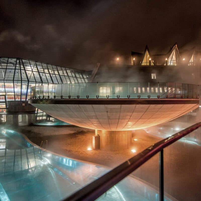 Futuristic building with a glass structure and a large bowl-shaped centerpiece resembling a thermal spa, illuminated by lights, surrounded by mist at night.