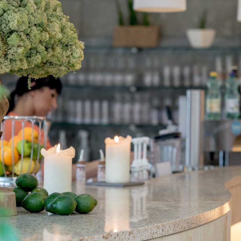 A calming thermal spa vibe envelops the scene, where a countertop adorned with candles, limes, and a vase of flowers completes the tranquil setting. In the background, someone is busy near shelves filled with bottles and containers.