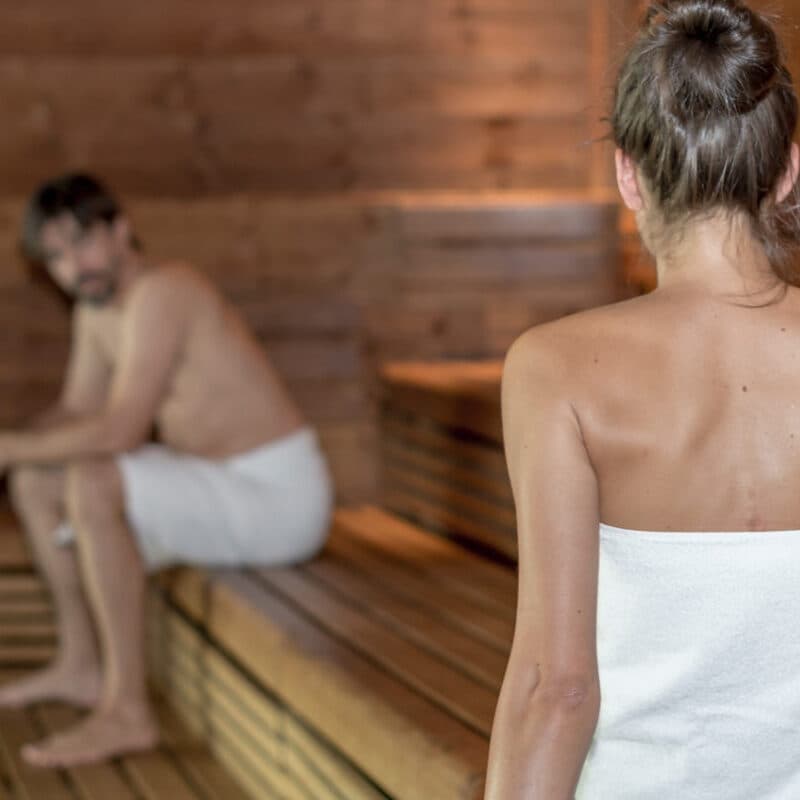 Two people wearing towels relax in a cozy wooden sauna, reminiscent of a serene thermal spa. One faces away from the camera while the other sits quietly on a bench in the background, absorbing the tranquil ambiance.