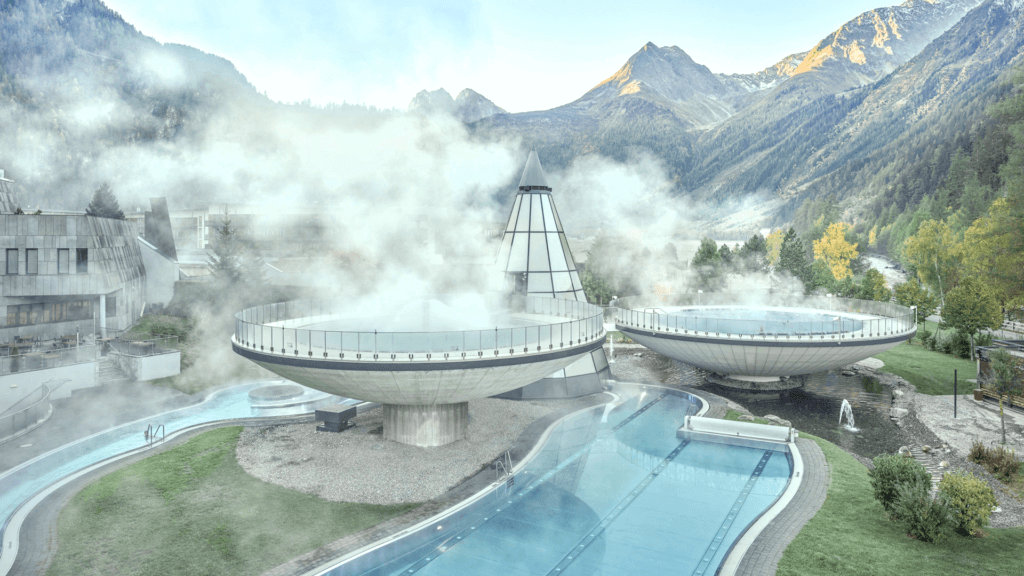 Edificio termale futuristico con strutture circolari che emettono vapore, circondato da montagne e alberi, con una lunga piscina all'aperto in primo piano.
