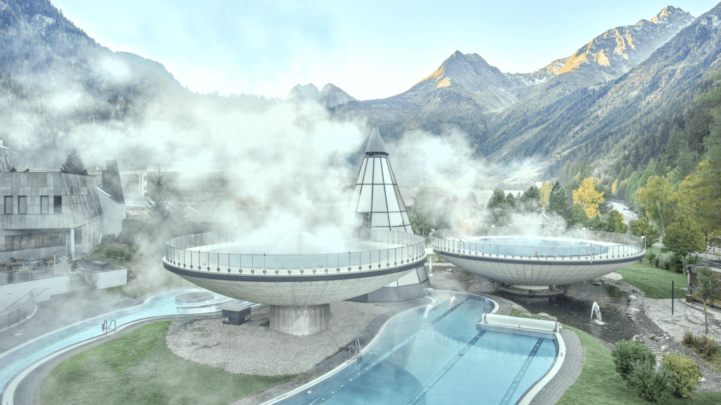 Vor einer Bergkulisse mit klarem Himmel stehen zwei kreisförmige Strukturen, aus denen Dampf austritt. Im Vordergrund sind außerdem ein dreieckiges Gebäude und ein gewundener Pool zu sehen.