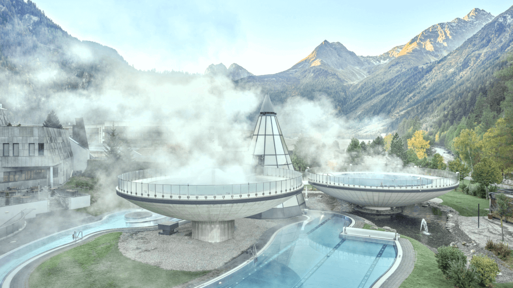 Spa extérieur avec vapeur s'élevant de piscines circulaires, entouré de montagnes et d'arbres.