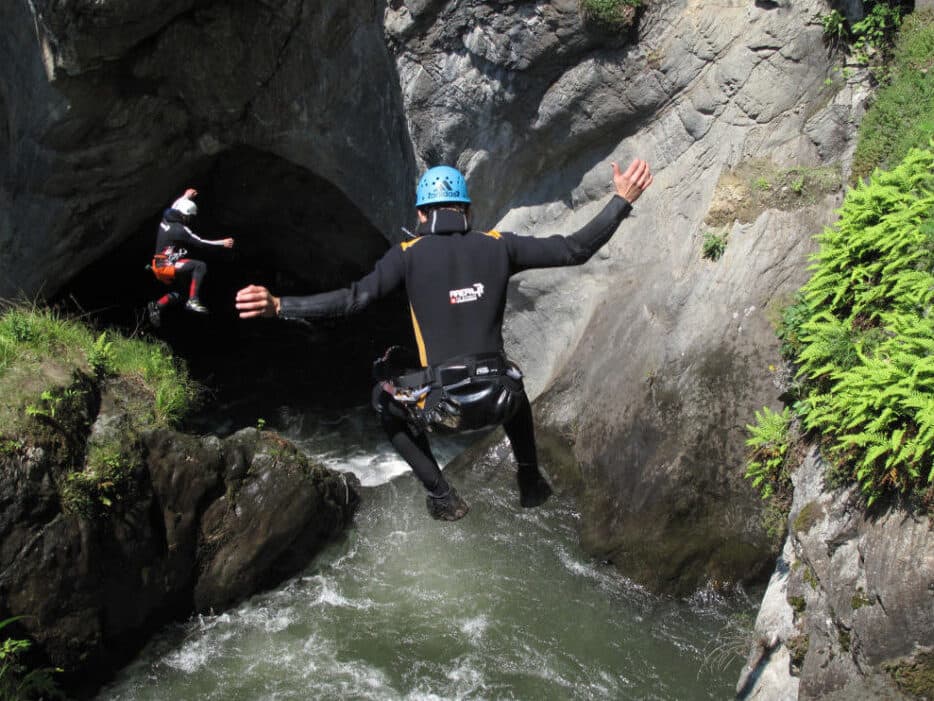Deux personnes, en combinaison et casque, se jettent dans une gorge rocheuse, où l'eau vive procure une poussée d'adrénaline digne d'une conférence palpitante. L'un des aventuriers est en plein vol, tandis que l'autre s'approche d'une arche, entourée d'une végétation luxuriante.