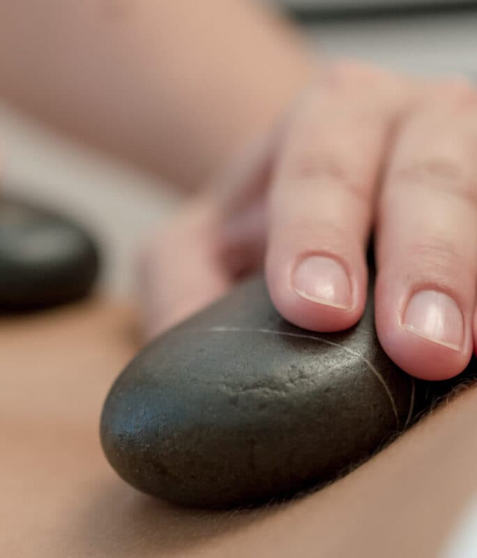 Nahaufnahme der Hand einer Person, die während einer Hot-Stone-Massage einen glatten, schwarzen Stein auf den Rücken einer Person legt.