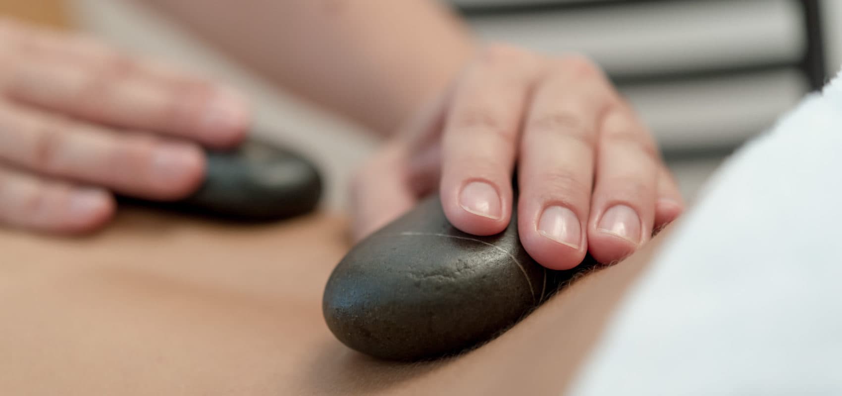 Person erhält eine Hot-Stone-Massage. Hände legen glatte, dunkle Steine auf den Rücken.