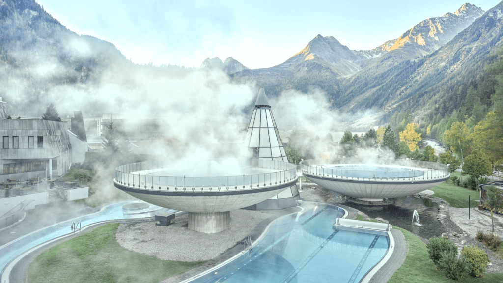 Futuristischer Spa-Komplex mit kreisförmigen, dampfenden Strukturen, umgeben von einem Pool. Gelegen in einer Bergregion mit Bäumen und Gipfeln im Hintergrund.
