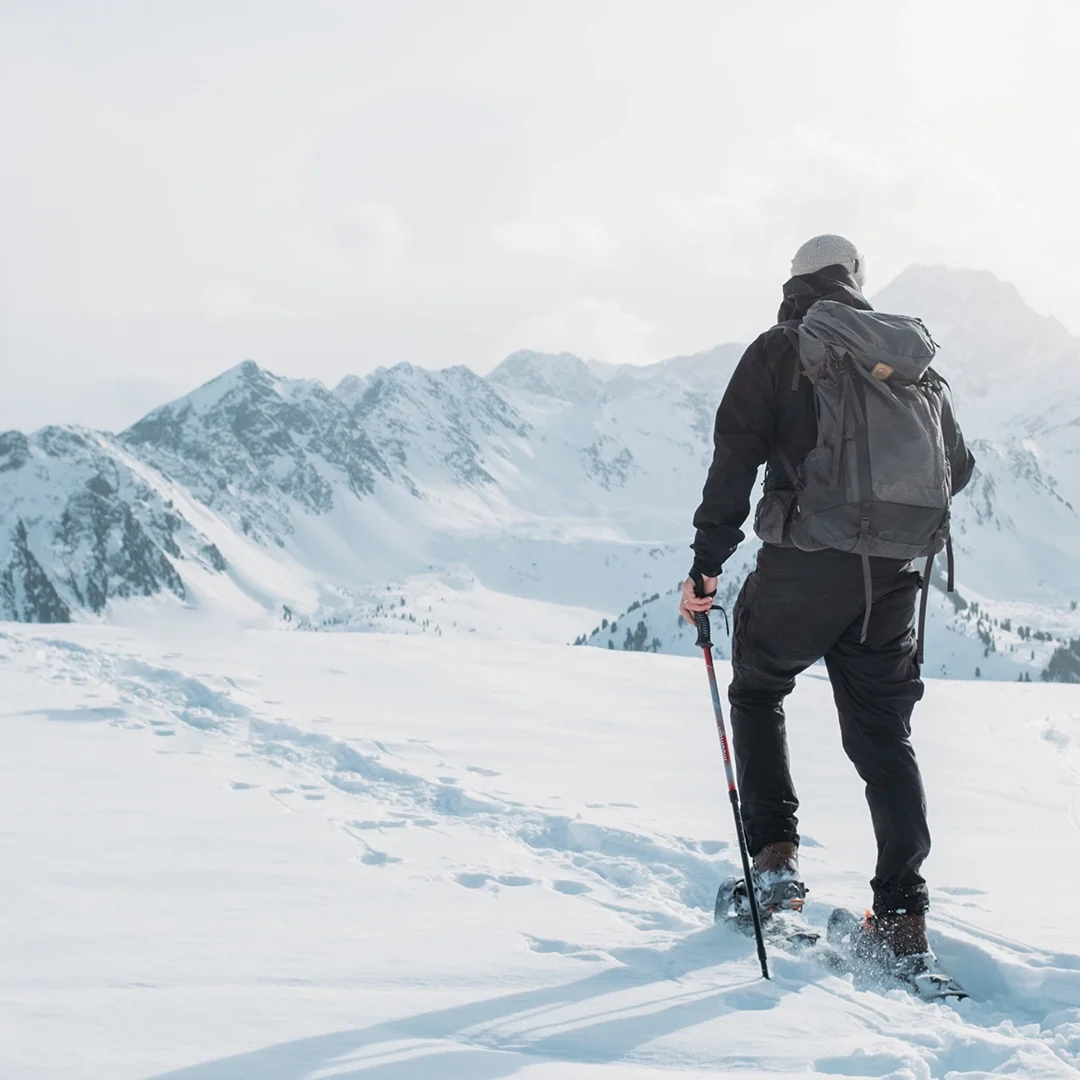 Person, die mit einem Wanderstock und einem Rucksack sowie Winterkleidung in einer verschneiten Berglandschaft mit Schneeschuhen wandert, im Hintergrund schneebedeckte Gipfel unter einem bewölkten Himmel.