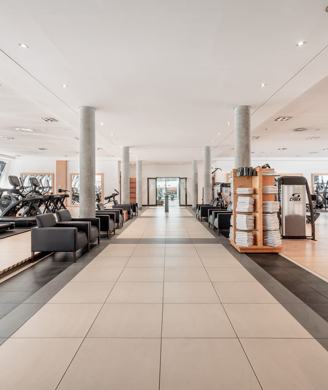 Großzügiger Fitnessraum mit Trainingsgeräten auf beiden Seiten, Säulen, Liegestühlen und einem Regal mit Handtüchern. Der Boden ist mit großen hellen Fliesen ausgelegt.