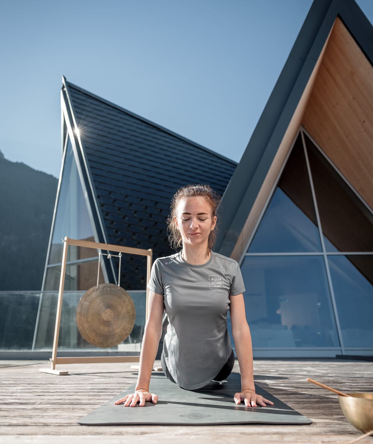 Una persona pratica yoga all'aperto su una terrazza di legno, abbracciando il suo percorso di fitness nella posizione del cane a testa in su, con edifici dal tetto triangolare e un gong sullo sfondo.