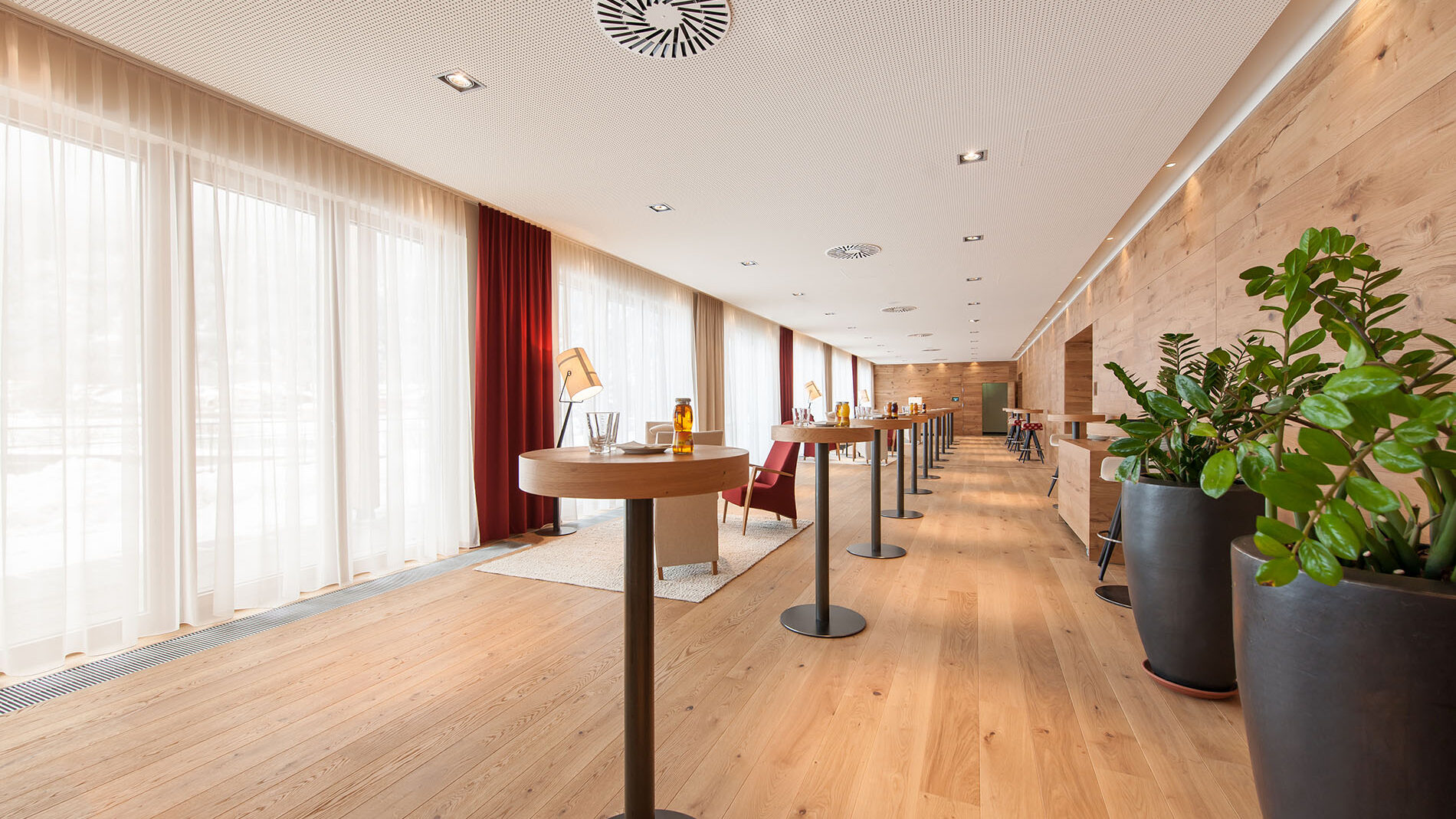 Der seminartaugliche Konferenzraum verfügt über hohe Tische, Holzboden und große Fenster mit roten Vorhängen. Topfpflanzen säumen die rechte Wand und sorgen für einen Hauch von Natur.