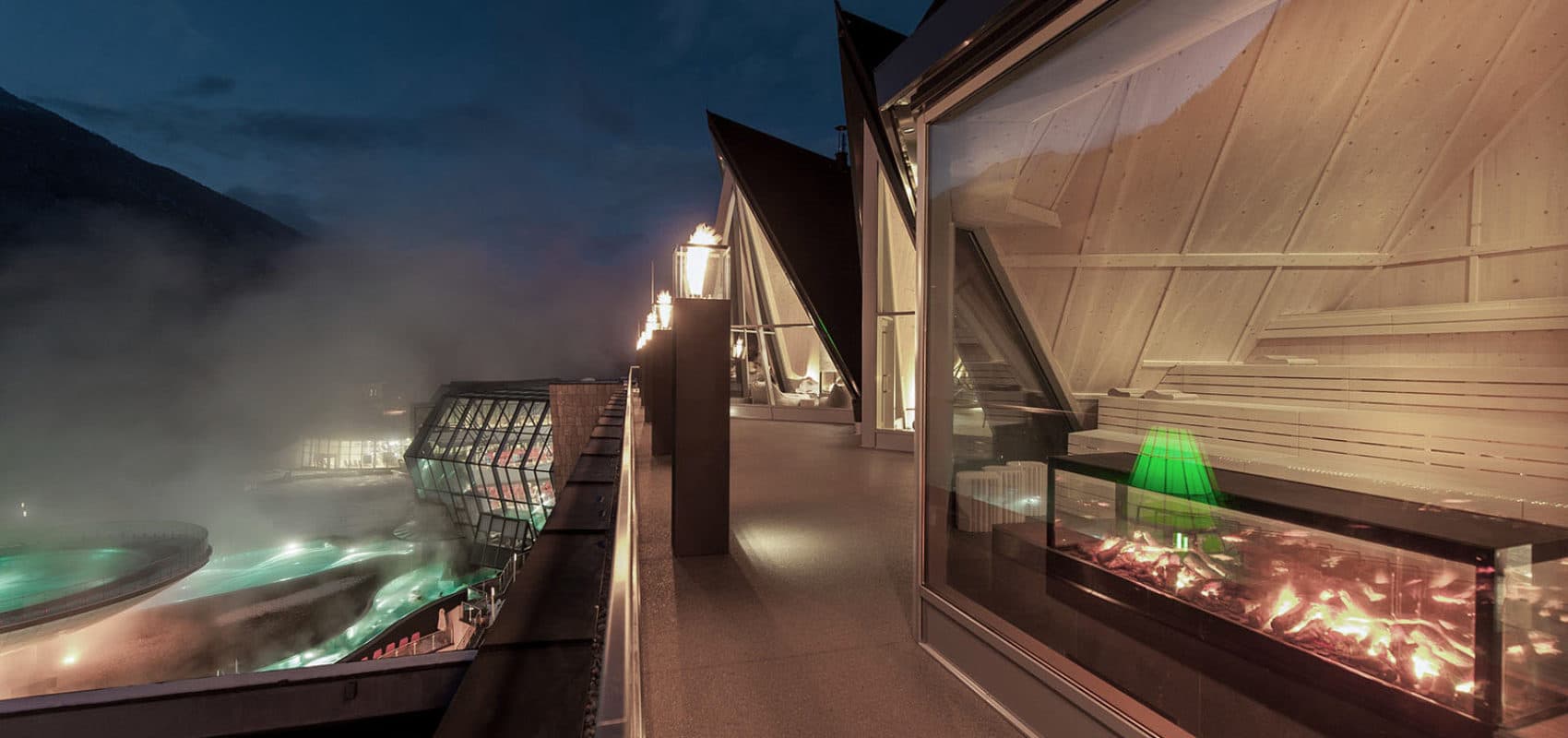 Exterior view of a modern building with large windows overlooking a foggy night scene. Lit interiors reveal wooden paneling, while an outdoor area displays illuminated water features.