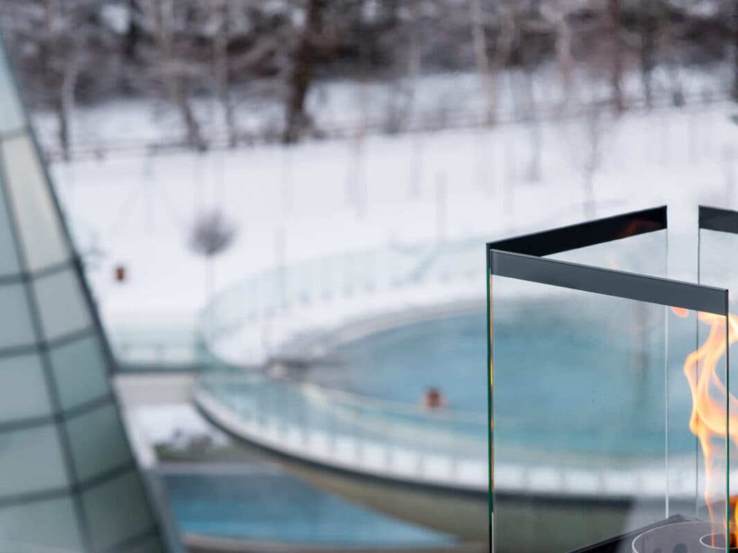 Nahaufnahme eines modernen Glaskamins mit sichtbarer Flamme und Blick auf eine schneebedeckte Landschaft und einen runden Pool.